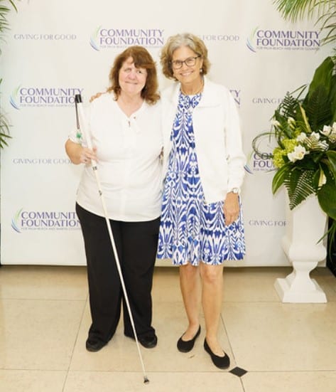 woman in shite cane stands next to woman in blue dress in front of a Community Foundation step and repeat