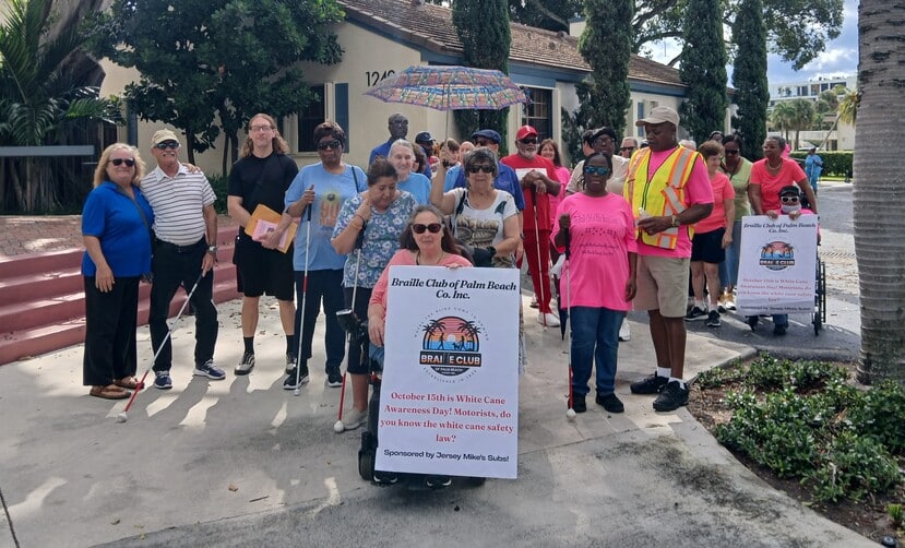 group of adults stand with a braille club sign while one woman holds an umbrella