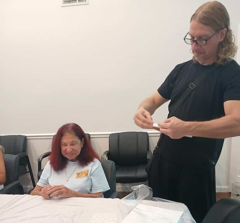 man wearing all black stands by table holding a canvas with woman sitting in background