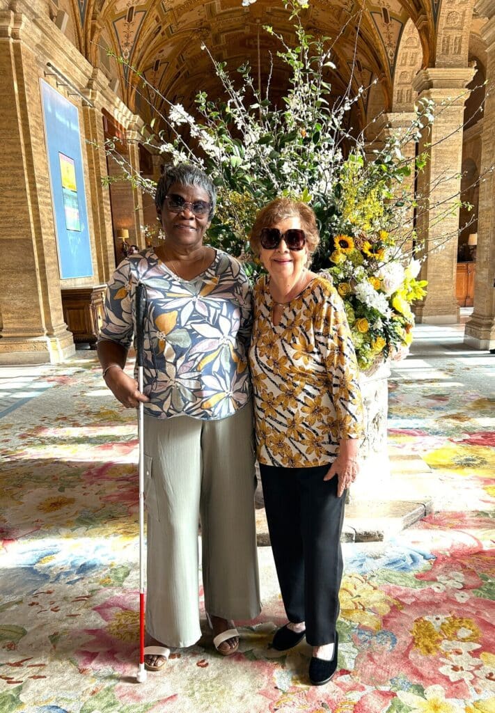 Woman in sunglasses and white cane stand with woman in sunglasses in front of a large flower arrangement in hotel lobby