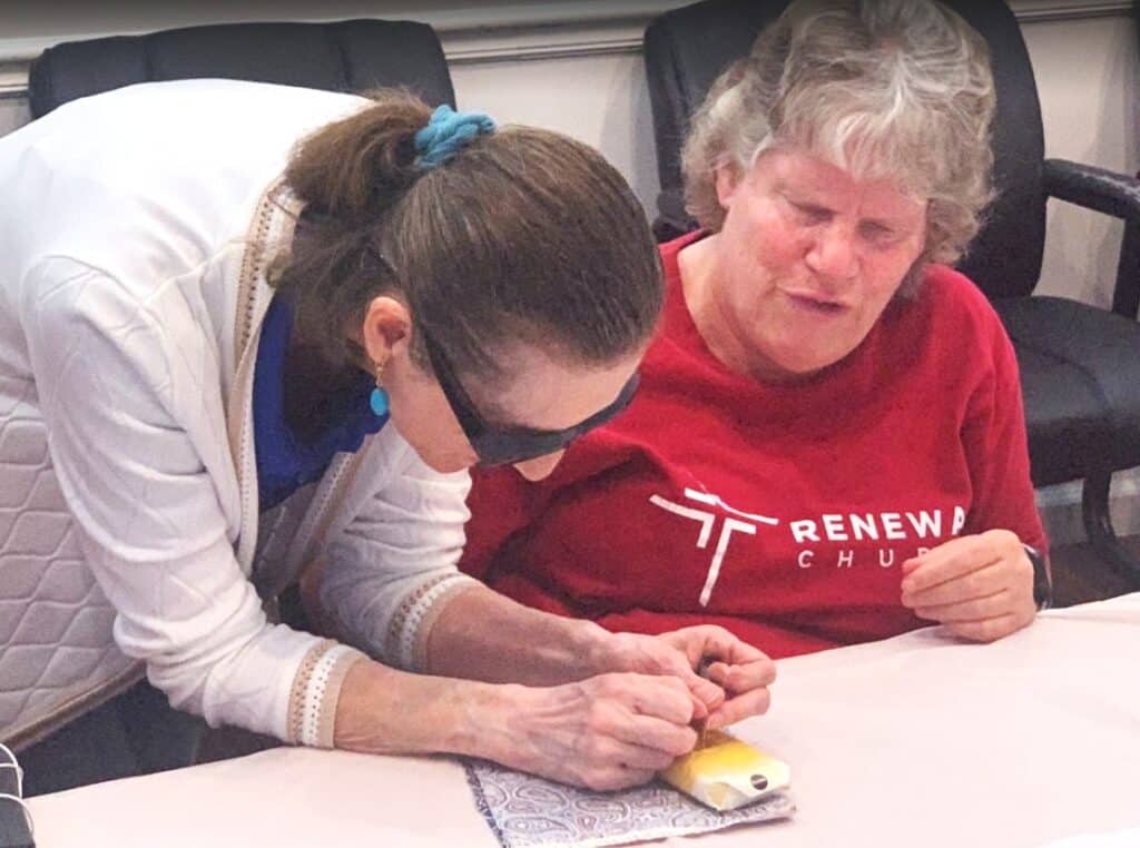 Woman in sunglasses bends over table to help woman in red shirt with craft