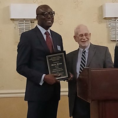 Man stands at podium holding an award next to another man
