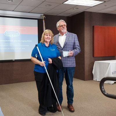 woman with white cane stands with man in suit jacket holding a check