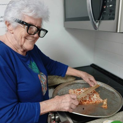 woman in blue shirt and two pairs of glasses on cuts pizza on tray in kitchen