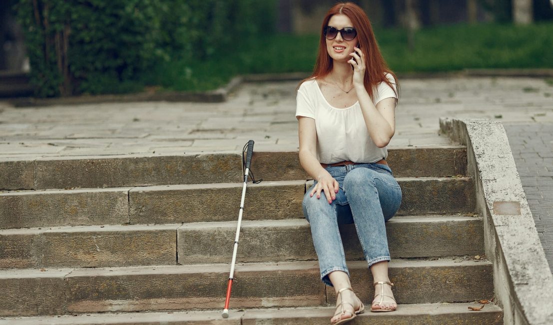 woman with white cane, sitting outside on steps and talking on the phone